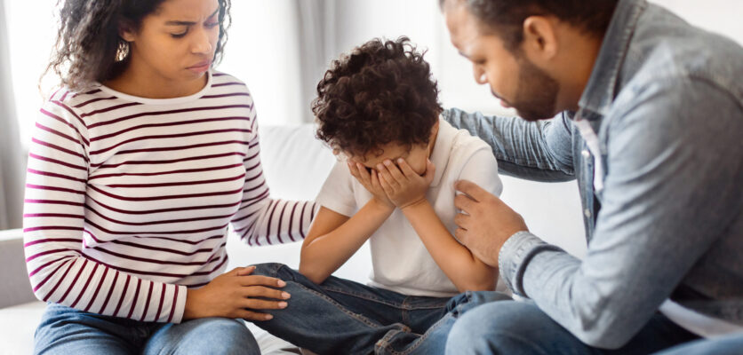 A Man and Woman Sitting on a Couch With Child African American man and woman are seated on a couch, accompanied by a child. Parents comforting their upset crying child, home interior