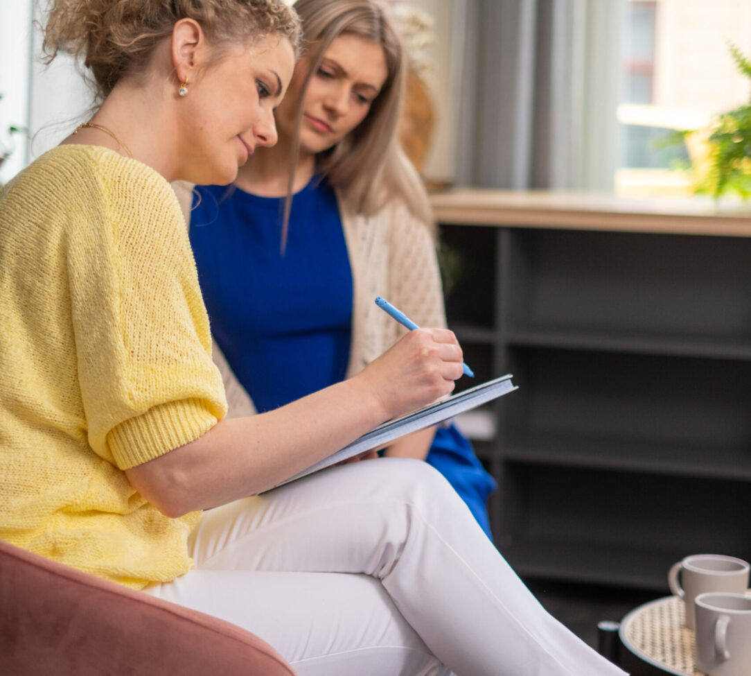 Two professional women engage in a coaching or mental therapy session, one taking notes, suggesting a focused approach to personal growth and business development. Description 2: Two professional women engage in a coaching or mental therapy session, one taking notes, suggesting a focused approach to personal growth and business development. Description 2:. High quality photo